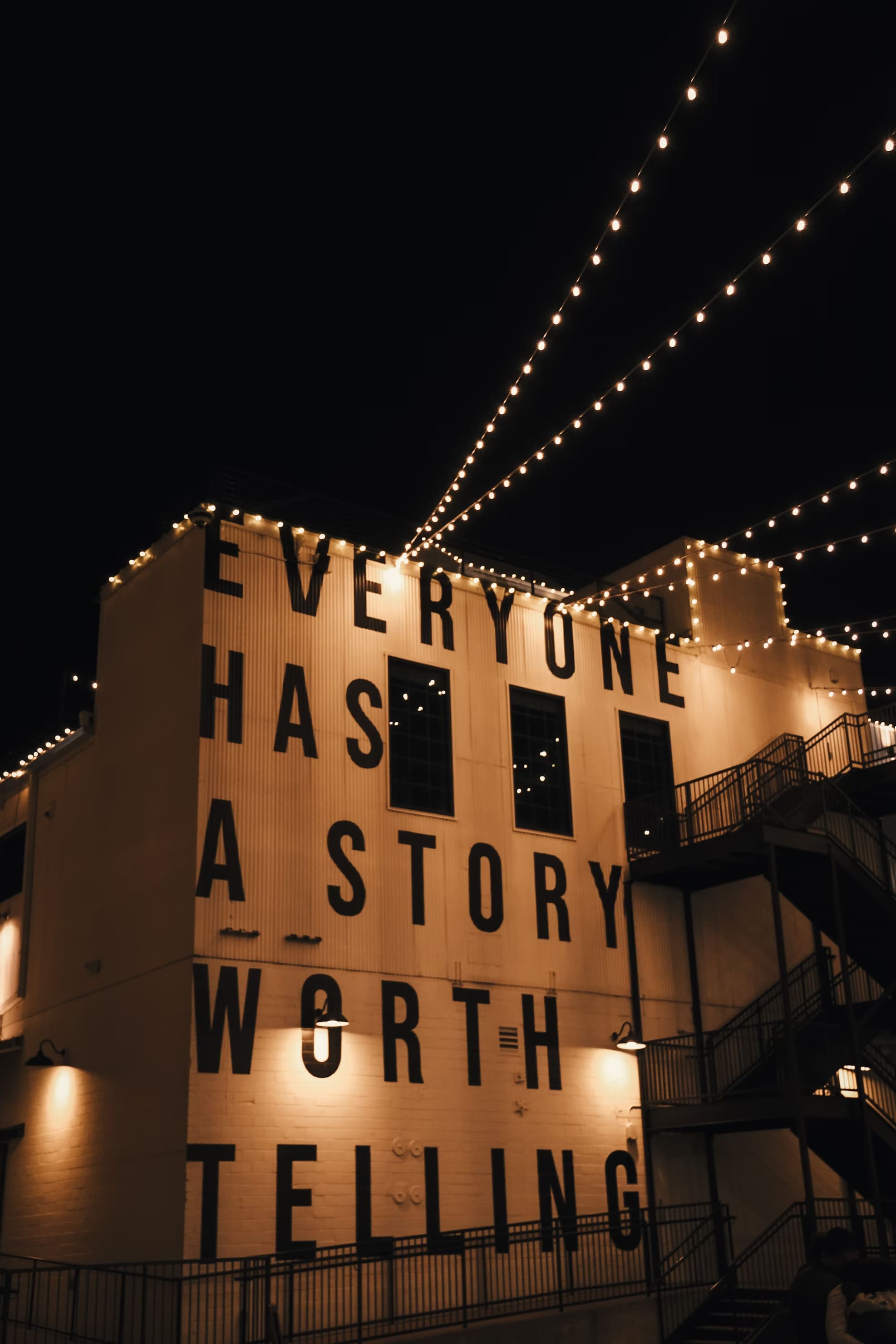 un immeuble la nuit illuminé par des guirlandes - couleurs chaudes. Sur la façade  est écrit : every one has a story worth telling (tout le monde à une histoire qui mérite d'être racontée
