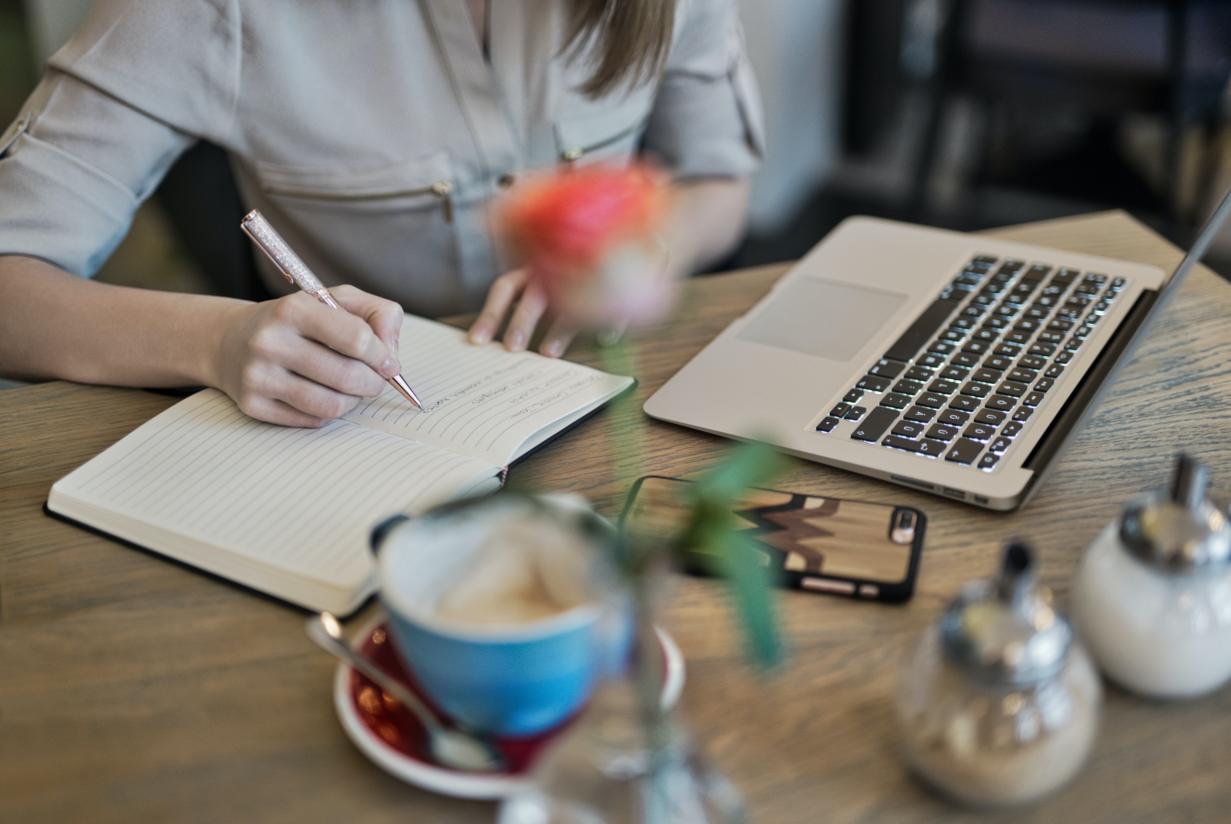 buste de femme devant un bureau. elle écrit sur un carnet. ordinateur portable devant elle et rose floue dans un vase, en premier plan